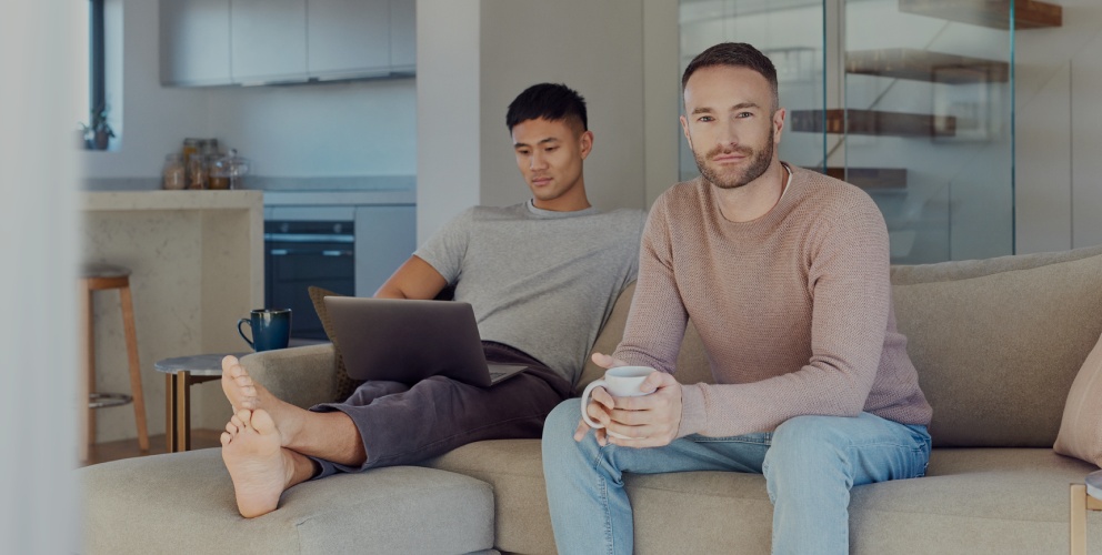 Zwei Männer sitzen zu Hause auf einem Sofa. Einer hat einen Laptop auf dem Schoß, der andere schaut freundlich in die Kamera.