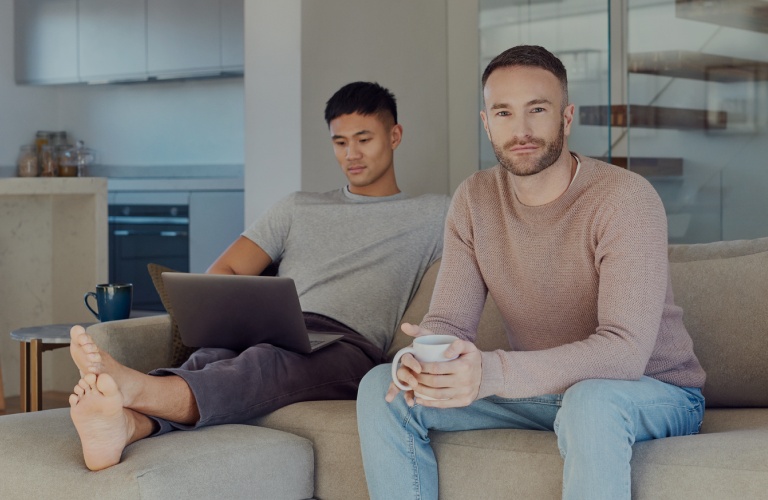 Zwei Männer sitzen zu Hause auf einem Sofa. Einer hat einen Laptop auf dem Schoß, der andere schaut freundlich in die Kamera.