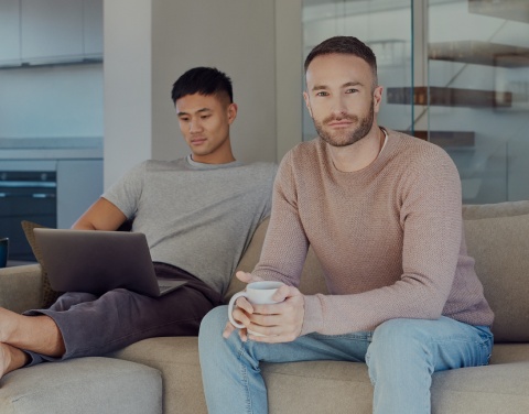 Zwei Männer sitzen zu Hause auf einem Sofa. Einer hat einen Laptop auf dem Schoß, der andere schaut freundlich in die Kamera.