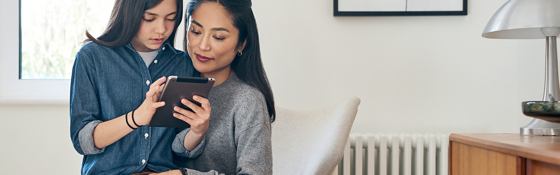 Ein Mädchen sitzt mit ihrem Tablet in der Hand auf dem Schoß ihrer Mutter und erklärt ihr etwas. Die Mutter hört gespannt zu. Beide befinden sich in einem hellen und modernen Raum.