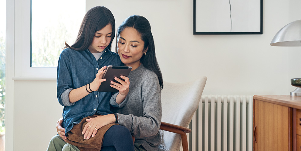 Ein Mädchen sitzt mit ihrem Tablet in der Hand auf dem Schoß ihrer Mutter und erklärt ihr etwas. Die Mutter hört gespannt zu. Beide befinden sich in einem hellen und modernen Raum.