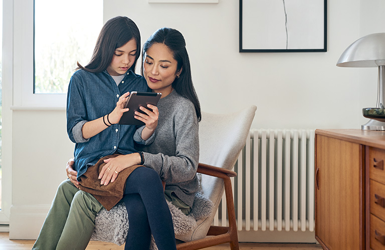 Ein Mädchen sitzt mit ihrem Tablet in der Hand auf dem Schoß ihrer Mutter und erklärt ihr etwas. Die Mutter hört gespannt zu. Beide befinden sich in einem hellen und modernen Raum.