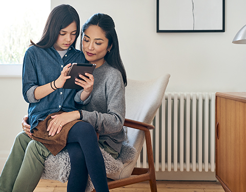 Ein Mädchen sitzt mit ihrem Tablet in der Hand auf dem Schoß ihrer Mutter und erklärt ihr etwas. Die Mutter hört gespannt zu. Beide befinden sich in einem hellen und modernen Raum.