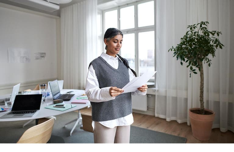 Eine Frau steht in einem hellen und modernen Arbeitszimmer. Sie hält mehrere Zettel in der Hand und liest diese aufmerksam durch.
