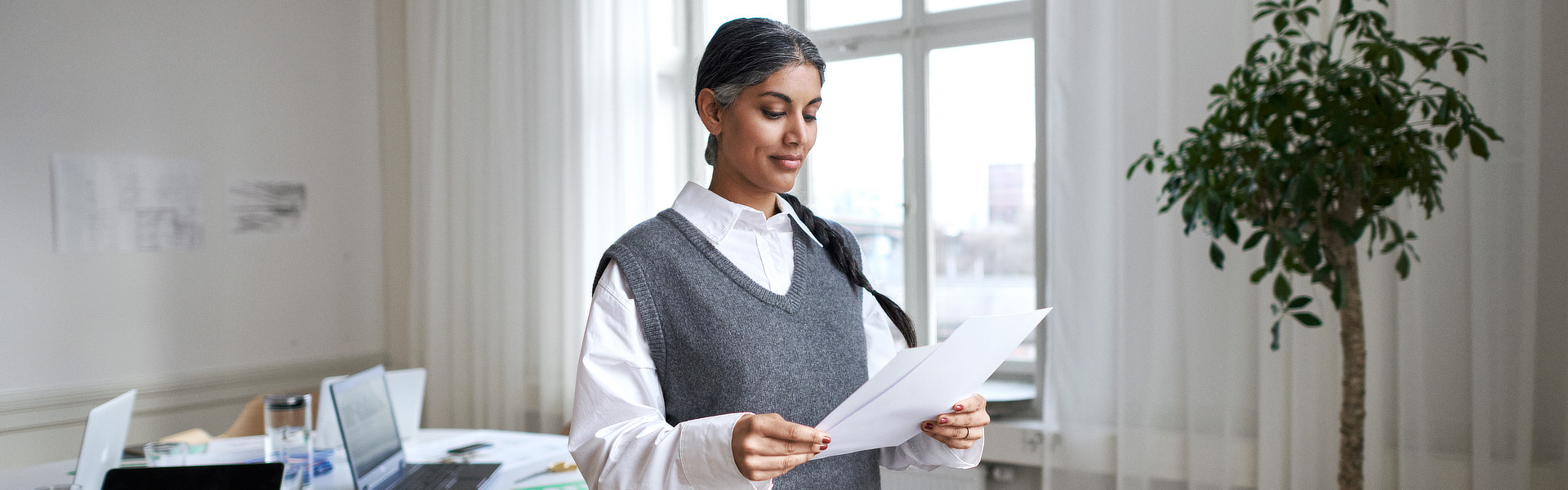 Eine Frau steht in einem hellen und modernen Arbeitszimmer. Sie hält mehrere Zettel in der Hand und liest diese aufmerksam durch.