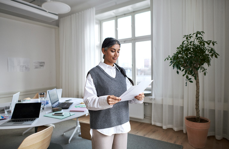 Eine Frau steht in einem hellen und modernen Arbeitszimmer. Sie hält mehrere Zettel in der Hand und liest diese aufmerksam durch.