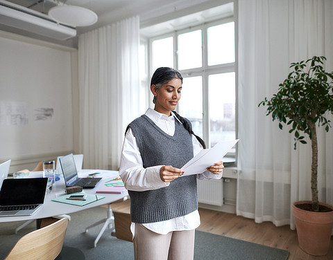 Eine Frau steht in einem hellen und modernen Arbeitszimmer. Sie hält mehrere Zettel in der Hand und liest diese aufmerksam durch.
