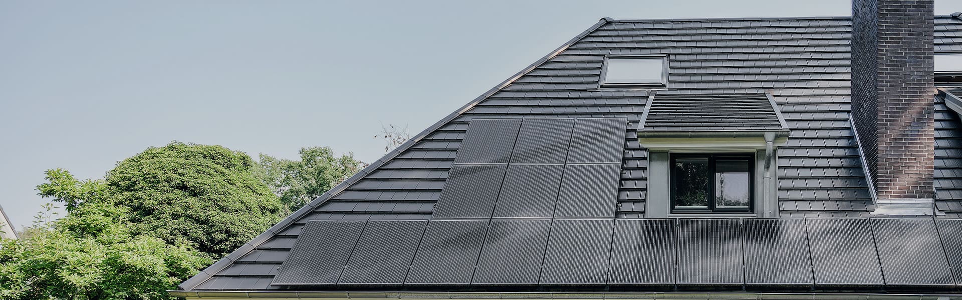 Ein schwarzes Hausdach mit integrierter Photovoltaikanlage im Vordergrund und grüne Bäume im Hintergrund.