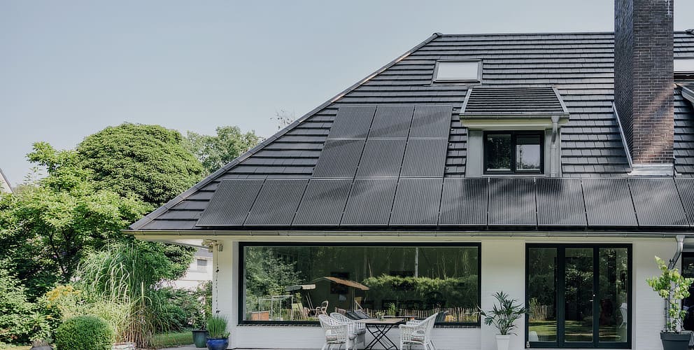 Ein schwarzes Hausdach mit integrierter Photovoltaikanlage im Vordergrund und grüne Bäume im Hintergrund.