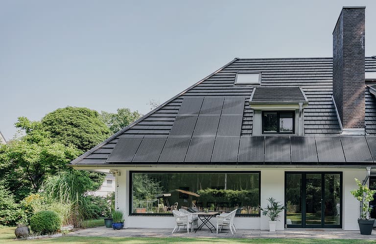 Ein schwarzes Hausdach mit integrierter Photovoltaikanlage im Vordergrund und grüne Bäume im Hintergrund.