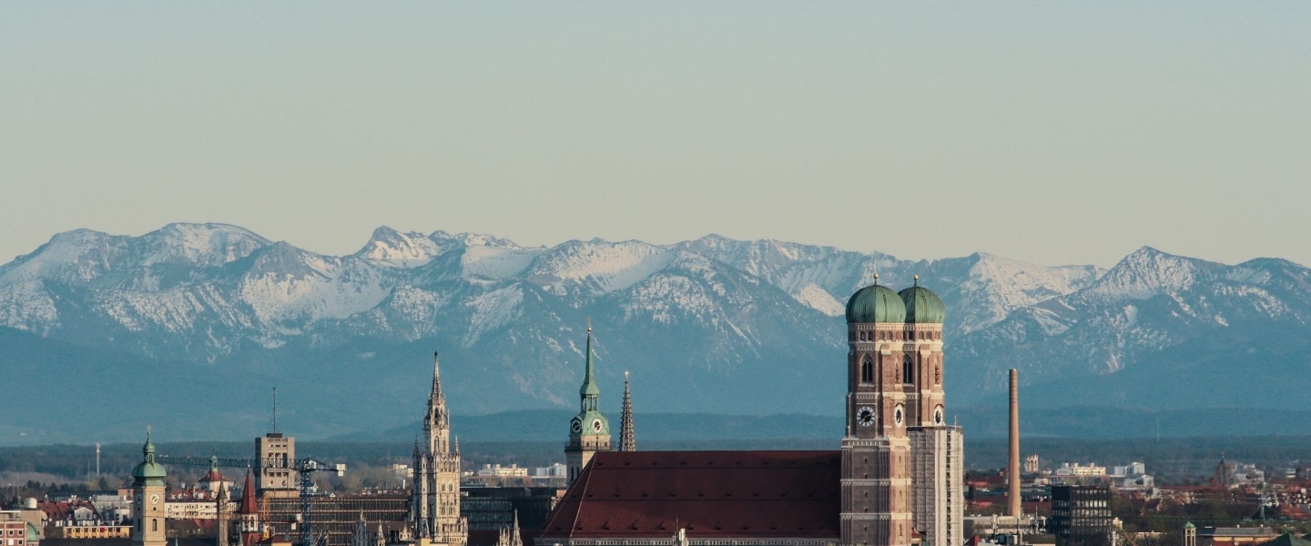 Blick über die Skyline von München mit der Frauenkirche im Vordergrund und den Bergen im Hintergrund.