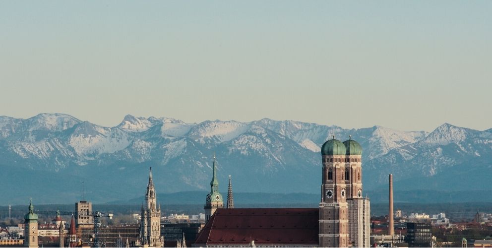 Blick über die Skyline von München mit der Frauenkirche im Vordergrund und den Bergen im Hintergrund.