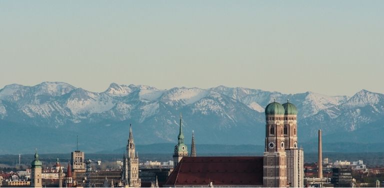 Blick über die Skyline von München mit der Frauenkirche im Vordergrund und den Bergen im Hintergrund.