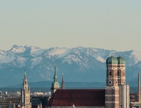 Blick über die Skyline von München mit der Frauenkirche im Vordergrund und den Bergen im Hintergrund.
