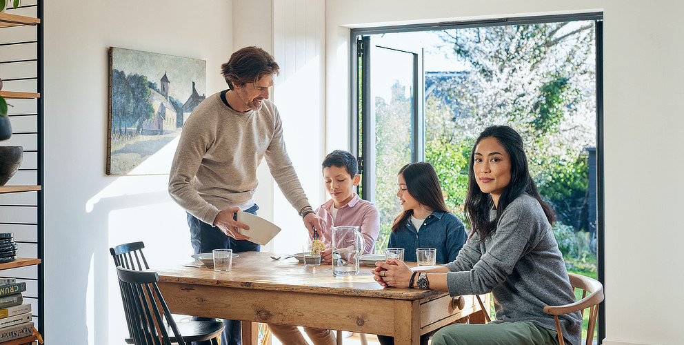 Eine Familie, bestehend aus einem Sohn, einer Tochter, einem Vater und einer Mutter, sitzt an einem Esstisch in einem modernen und hellen Raum mit einer offenen Terassentür aus Glas. Im Hintergrund sieht man einen Garten. Der Vater gibt seinem Sohn etwas zu Essen auf den Teller.