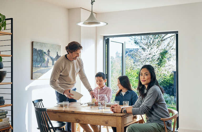 Eine Familie, bestehend aus einem Sohn, einer Tochter, einem Vater und einer Mutter, sitzt an einem Esstisch in einem modernen und hellen Raum mit einer offenen Terassentür aus Glas. Im Hintergrund sieht man einen Garten. Der Vater gibt seinem Sohn etwas zu Essen auf den Teller.