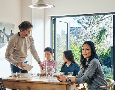Eine Familie, bestehend aus einem Sohn, einer Tochter, einem Vater und einer Mutter, sitzt an einem Esstisch in einem modernen und hellen Raum mit einer offenen Terassentür aus Glas. Im Hintergrund sieht man einen Garten. Der Vater gibt seinem Sohn etwas zu Essen auf den Teller.
