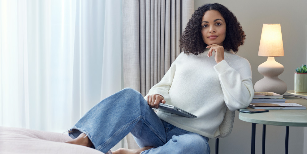 Eine Frau sitzt mit einem weißen, warmen Pullover in einem modernen hellen Raum. Im Anschnitt leuchtet im Hintergrund eine moderne Tischlampe.