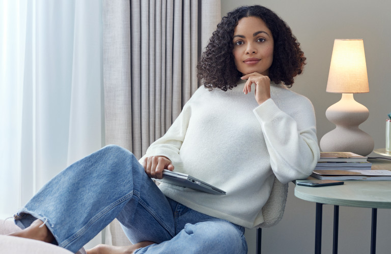 Eine Frau sitzt mit einem weißen, warmen Pullover in einem modernen hellen Raum. Im Anschnitt leuchtet im Hintergrund eine moderne Tischlampe.