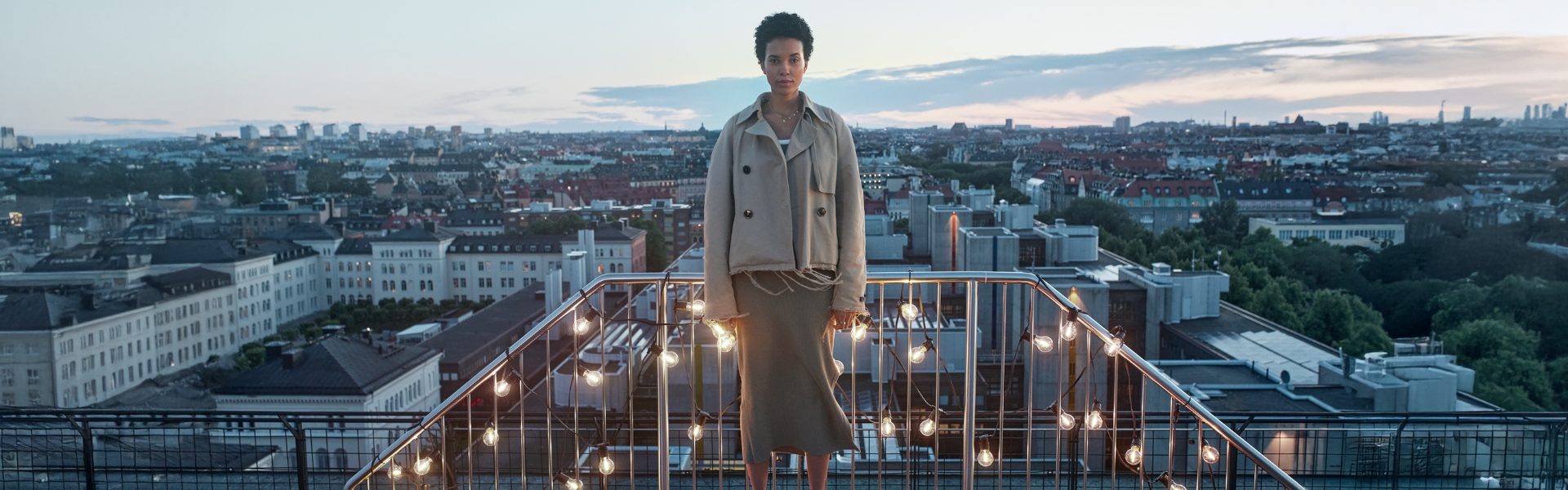 Eine Frau steht an einem Balkongeländer mit einer Lichterkette, die warmes Licht abgibt. Im Hintergrund sind die Dächer einer Stadt zusehen.