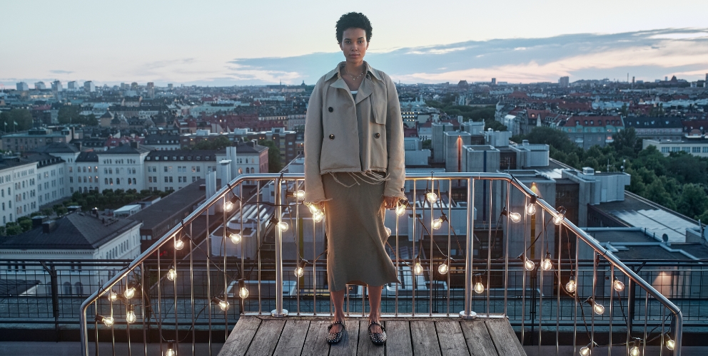 Eine Frau steht an einem Balkongeländer mit einer Lichterkette, die warmes Licht abgibt. Im Hintergrund sind die Dächer einer Stadt zusehen.