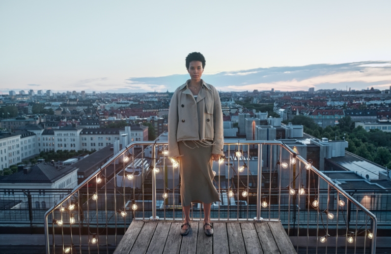 Eine Frau steht an einem Balkongeländer mit einer Lichterkette, die warmes Licht abgibt. Im Hintergrund sind die Dächer einer Stadt zusehen.
