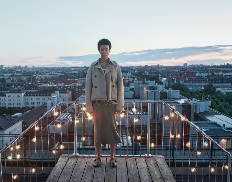 Eine Frau steht an einem Balkongeländer mit einer Lichterkette, die warmes Licht abgibt. Im Hintergrund sind die Dächer einer Stadt zusehen.