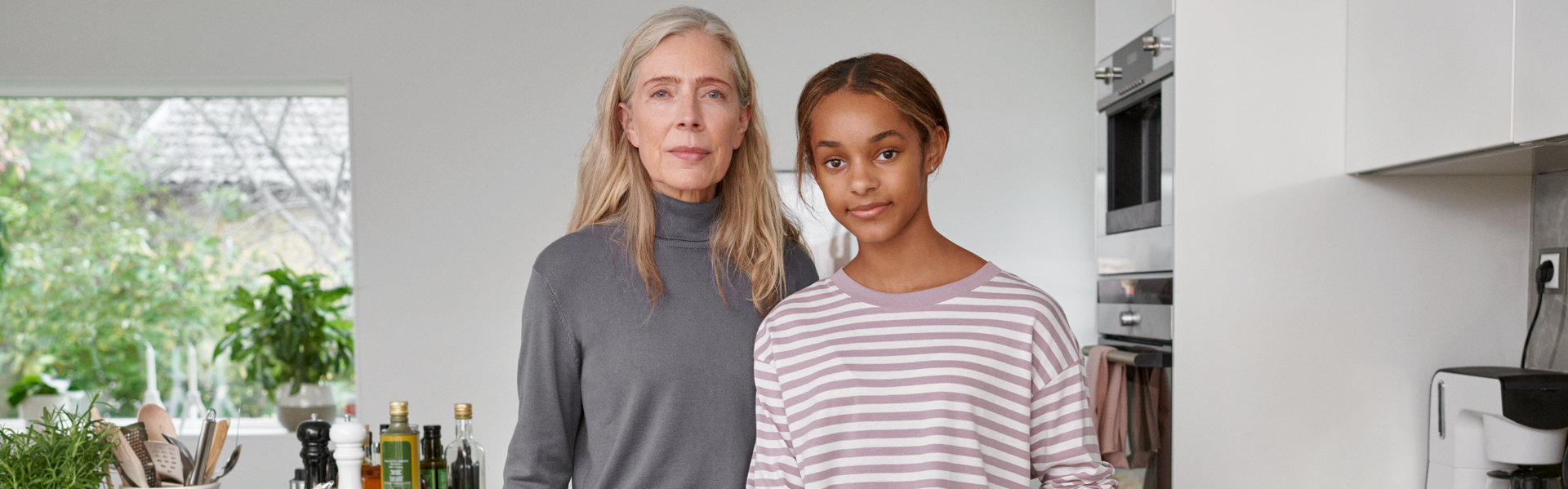 Eine Frau und ein Mädchen stehen eng nebeneinander in einer modernen und hellen Küche. Beide blicken ein wenig lächelnd direkt in die Kamera.
