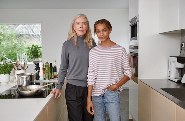 Eine Frau und ein Mädchen stehen eng nebeneinander in einer modernen und hellen Küche. Beide blicken ein wenig lächelnd direkt in die Kamera.