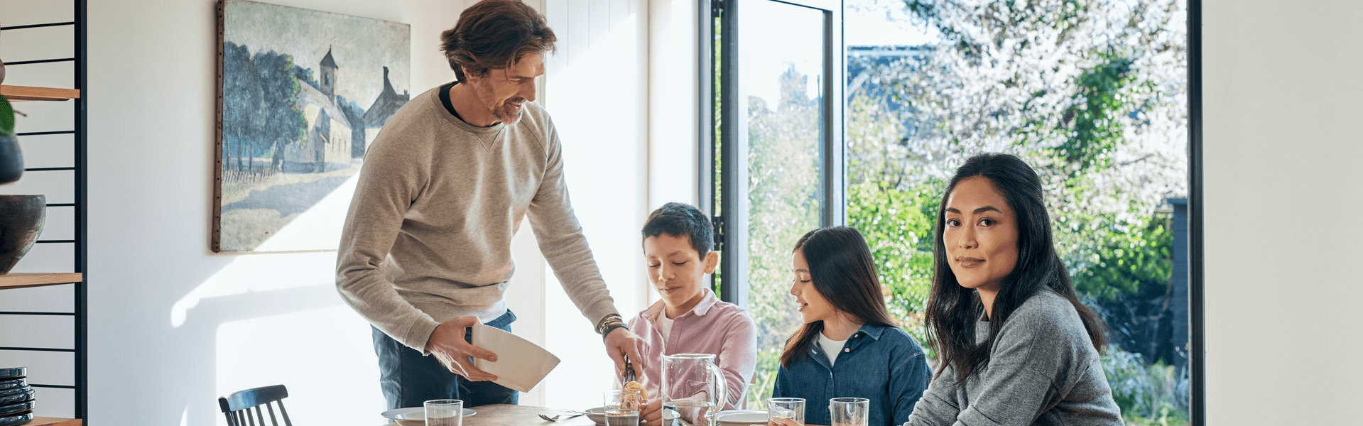 Eine Familie, bestehend aus einem Sohn, einer Tochter, einem Vater und einer Mutter, sitzt an einem Esstisch in einem modernen und hellen Raum mit einer offenen Terassentür aus Glas. Im Hintergrund sieht man einen Garten. Der Vater gibt seinem Sohn etwas zu Essen auf den Teller.