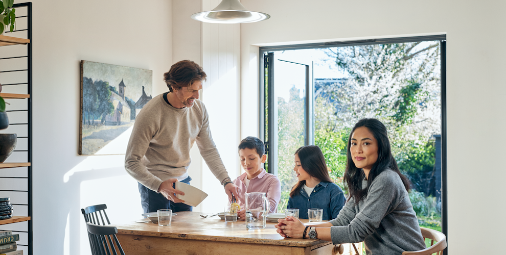 Eine Familie, bestehend aus einem Sohn, einer Tochter, einem Vater und einer Mutter, sitzt an einem Esstisch in einem modernen und hellen Raum mit einer offenen Terassentür aus Glas. Im Hintergrund sieht man einen Garten. Der Vater gibt seinem Sohn etwas zu Essen auf den Teller.