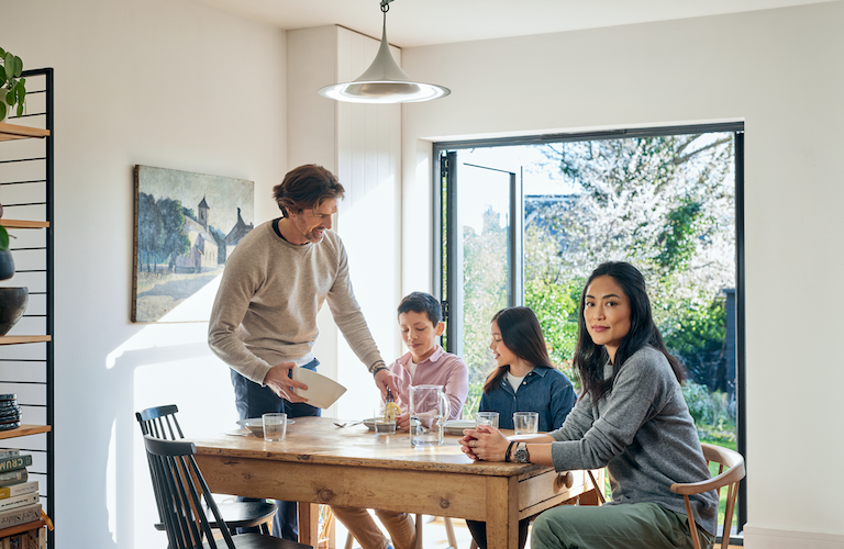 Eine Familie, bestehend aus einem Sohn, einer Tochter, einem Vater und einer Mutter, sitzt an einem Esstisch in einem modernen und hellen Raum mit einer offenen Terassentür aus Glas. Im Hintergrund sieht man einen Garten. Der Vater gibt seinem Sohn etwas zu Essen auf den Teller.