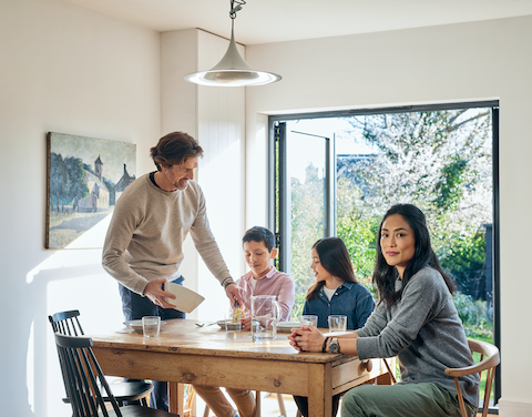 Eine Familie, bestehend aus einem Sohn, einer Tochter, einem Vater und einer Mutter, sitzt an einem Esstisch in einem modernen und hellen Raum mit einer offenen Terassentür aus Glas. Im Hintergrund sieht man einen Garten. Der Vater gibt seinem Sohn etwas zu Essen auf den Teller.