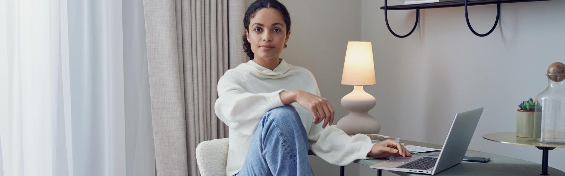 Eine Frau sitzt gemütlich vor ihrem Laptop an einem Tisch und schaut in die Kamera. Sie befindet sich in einem hellen und modernen Raum. Eine Lampe leuchtet im Hintergrund.