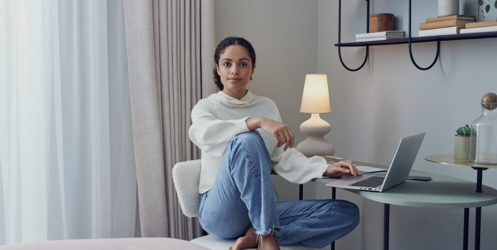 Eine Frau sitzt gemütlich vor ihrem Laptop an einem Tisch und schaut in die Kamera. Sie befindet sich in einem hellen und modernen Raum. Eine Lampe leuchtet im Hintergrund.