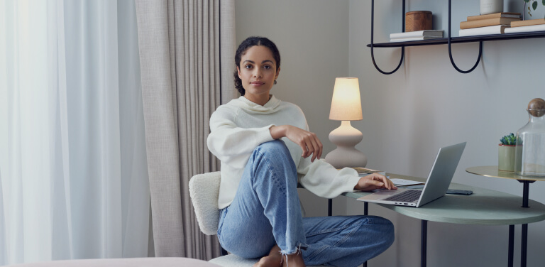 Eine Frau sitzt gemütlich vor ihrem Laptop an einem Tisch und schaut in die Kamera. Sie befindet sich in einem hellen und modernen Raum. Eine Lampe leuchtet im Hintergrund.