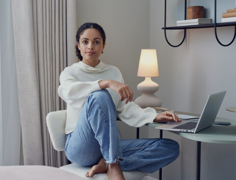Eine Frau sitzt gemütlich vor ihrem Laptop an einem Tisch und schaut in die Kamera. Sie befindet sich in einem hellen und modernen Raum. Eine Lampe leuchtet im Hintergrund.