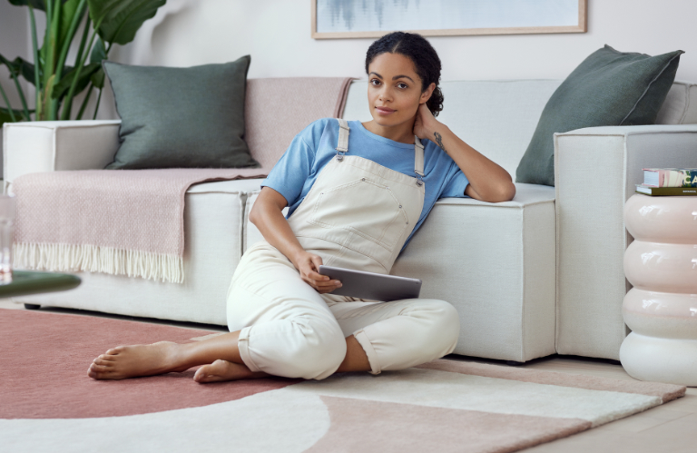Eine Frau sitzt auf dem Boden neben ihrem Sofa. Sie hat ein Tablet auf dem Schoß und schaut freundlich in die Kamera.