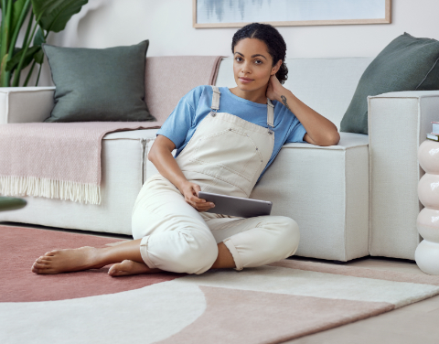 Eine Frau sitzt auf dem Boden neben ihrem Sofa. Sie hat ein Tablet auf dem Schoß und schaut freundlich in die Kamera.