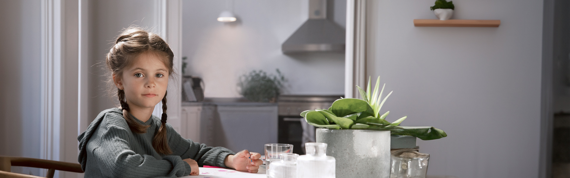 Ein Mädchen sitzt an einem Tisch in einer hellen Küche und malt mit Buntstiften. In der Küchenzeile leuchtet eine Lampe.