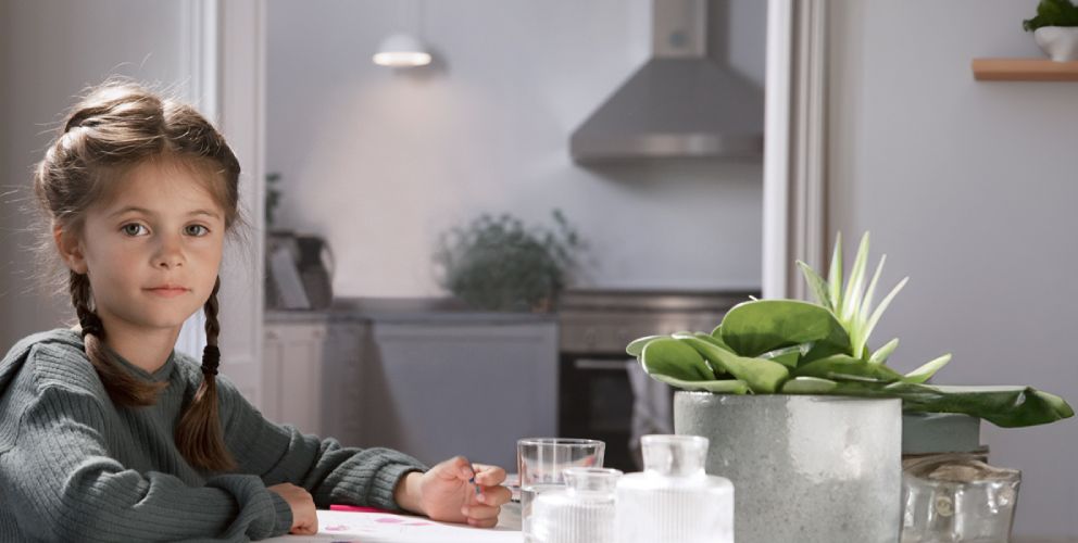 Ein Mädchen sitzt an einem Tisch in einer hellen Küche und malt mit Buntstiften. In der Küchenzeile leuchtet eine Lampe.