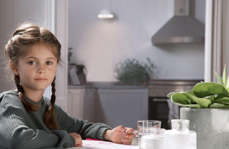 Ein Mädchen sitzt an einem Tisch in einer hellen Küche und malt mit Buntstiften. In der Küchenzeile leuchtet eine Lampe.
