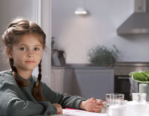 Ein Mädchen sitzt an einem Tisch in einer hellen Küche und malt mit Buntstiften. In der Küchenzeile leuchtet eine Lampe.