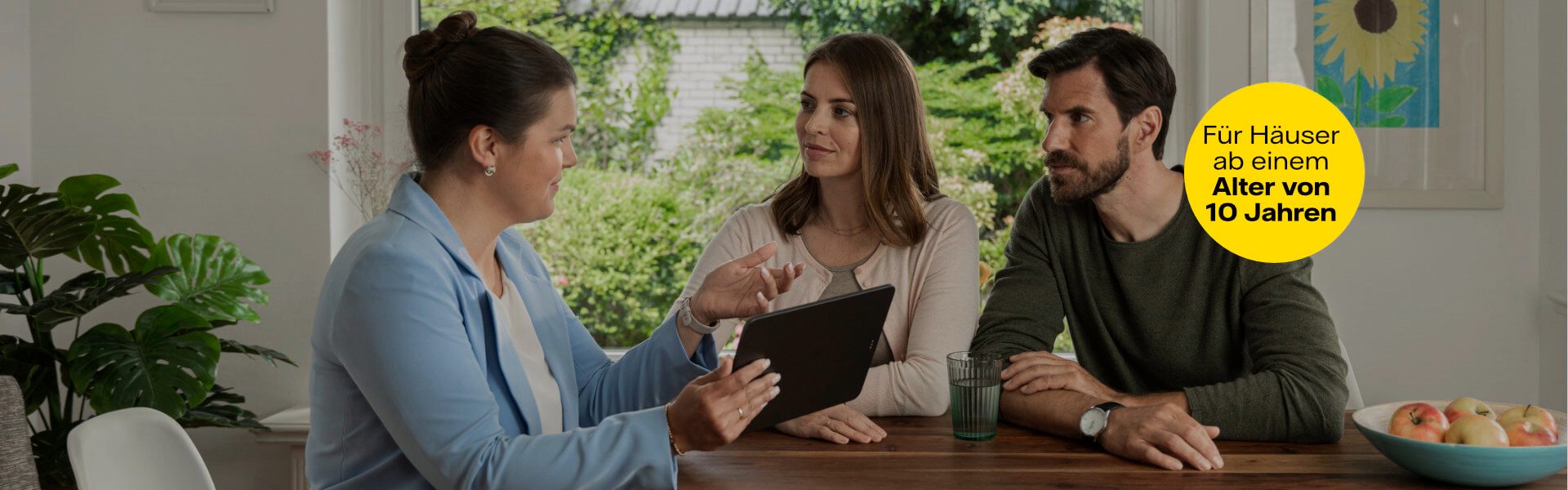 Eine Frau mit einem hellblauen Blazer sitzt gemeinsam mit einem Mann und einer Frau an einem Esstisch. Sie hält ein Tablet in der Hand und erklärt den beiden etwas. Sie befinden sich in einem modernen und hellen Esszimmer.