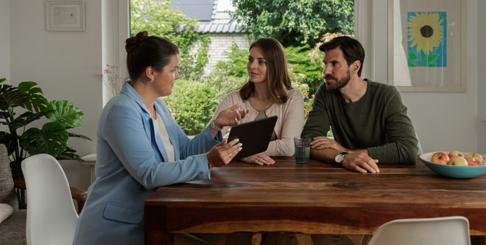Eine Frau mit einem hellblauen Blazer sitzt gemeinsam mit einem Mann und einer Frau an einem Esstisch. Sie hält ein Tablet in der Hand und erklärt den beiden etwas. Sie befinden sich in einem modernen und hellen Esszimmer.