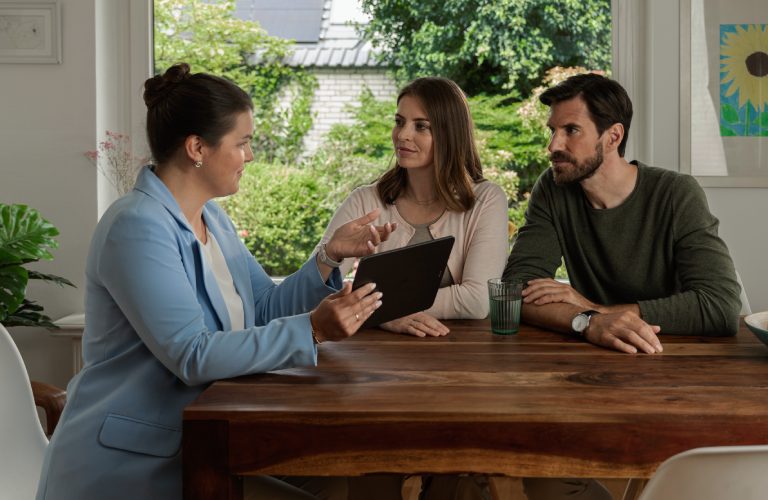 Eine Frau mit einem hellblauen Blazer sitzt gemeinsam mit einem Mann und einer Frau an einem Esstisch. Sie hält ein Tablet in der Hand und erklärt den beiden etwas. Sie befinden sich in einem modernen und hellen Esszimmer.