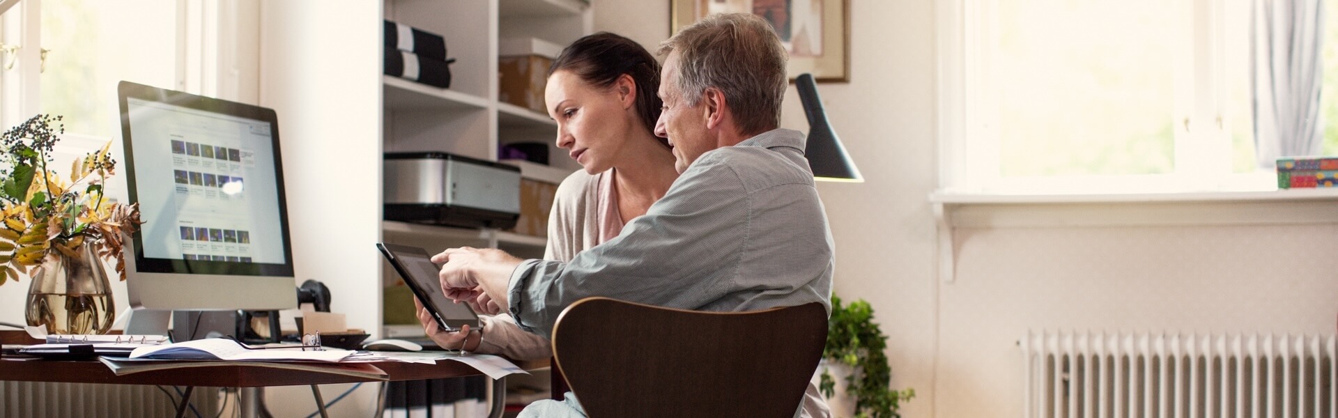 Mann und Frau sitzen in einem modernen, hellen Arbeitszimmer. Die Frau hält ein Tablet in der Hand und der Mann erklärt ihr etwas darauf. Sie hört gespannt zu.