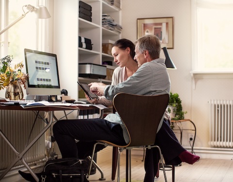 Mann und Frau sitzen in einem modernen, hellen Arbeitszimmer. Die Frau hält ein Tablet in der Hand und der Mann erklärt ihr etwas darauf. Sie hört gespannt zu.