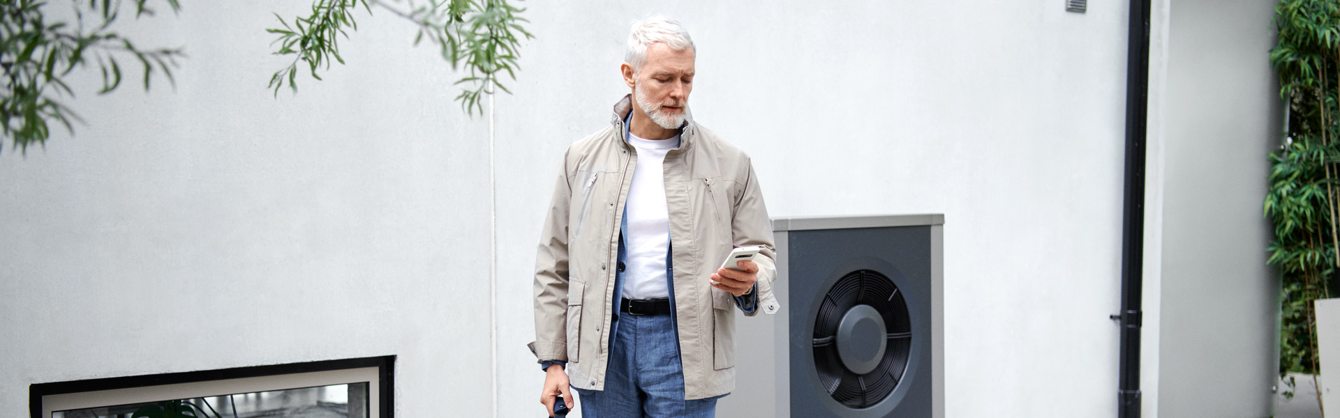 Ein Mann blickt auf sein Smartphone in seiner linken Hand und steht vor einer weißen Hauswand. Im Hintergrund ist eine moderne Wärmepumpe zu erkennen.