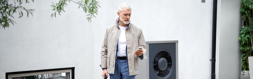 Ein Mann blickt auf sein Smartphone in seiner linken Hand und steht vor einer weißen Hauswand. Im Hintergrund ist eine moderne Wärmepumpe zu erkennen.